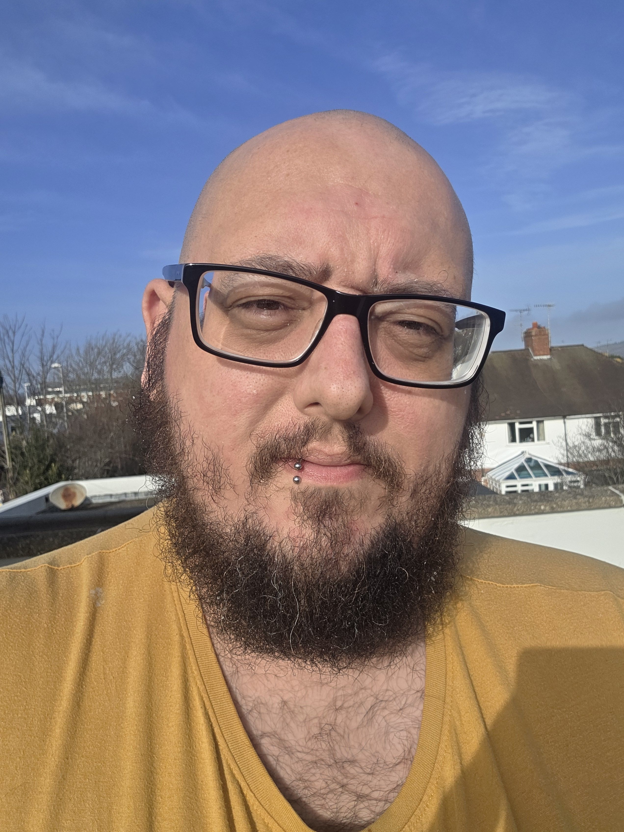 A close-up portrait of a bearded man with a shaved head, wearing glasses and a yellow shirt, standing outdoors in bright sunlight against a clear blue sky.